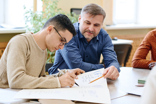 Mature Caucasian Teacher Of English Language Watching Middle Eastern Immigrant Student Writing Something On Poster