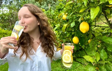 it's just a lemon paradise, a teenage girl stands against the backdrop of lemon tree with lemons and drinks lemonade from a glass glass in her other hand, she holds a jug of drink. High quality photo