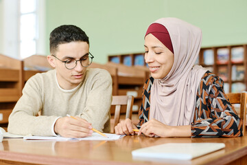 Young man and woman in hijab sitting at desk in classroom having lesson for immigrants working on...