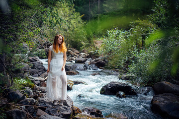 Slender long-haired brunette in white dress posing against small mountain river and green trees. Beautiful young woman walking along the forest stream shore.