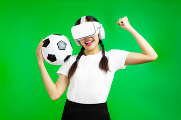 Cheerful asian girl football fan in vr glasses holding a soccer ball isolated green wall...
