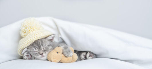 Fototapeta na wymiar Cozy tiny kitten wearing warm hat sleeps with favorite toy bear under warm white blanket on a bed at home. Empty space for text