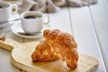 A crispy croissant with almond filling and hot black coffee on a wooden board on a light concrete table.