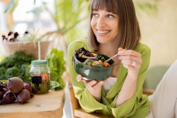 Young cheerful woman eats vegetarian lunch in bowl, sitting by the table full of fresh food...