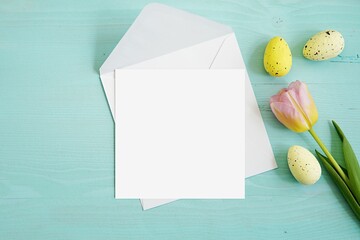 Square Easter greeting card mockup with white envelope for design or art presentation, flat lay composition on light blue table with decorative Easter eggs and tulip.