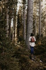 Obraz premium Hiking in the woods. A woman is standing in the forest, carrying leather backpack and watching the tall trees. Autumn activity in Slovenia, Europe. Pinewood trees, sunlight. 