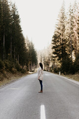 A woman is standing on an empty street in the forest and watching the trees. She is in the middle of the road. Blurry and hazy background, large pine trees. Woodland outdoor traveling. 