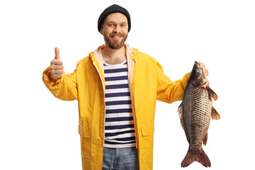 Young fisherman in a yellow raincoat holding a carp fish and gesturing thumbs up