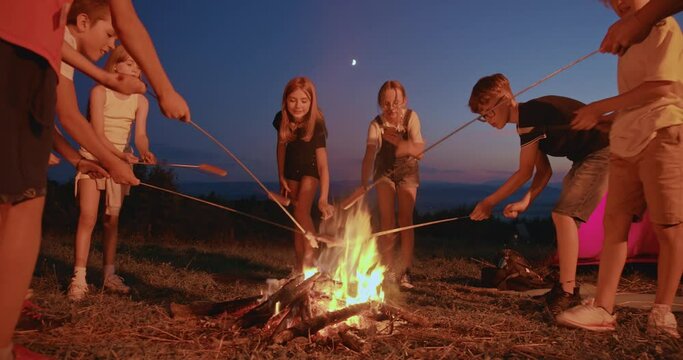Side view of kids raising up, frying sausages in fireplace in summer. Children hiking, having dinner in mountains near red tent. Concept of traveling, healthy lifestyle.