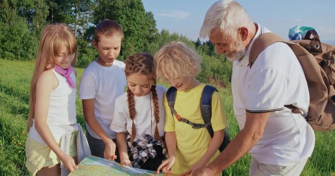 Front view of travelers holding map together in mountains. Four children standing, looking, showing, pointing, old man smiling. Concept of tourism and nature discovering.