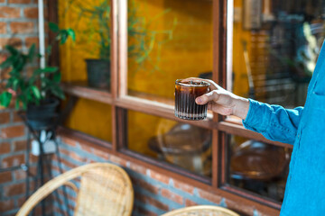 Obraz premium Hand holding a glass of ice coffee, americano in the afternoon over a cafe glass window background at a Cafe coffee shop outdoor. Cold brew refreshment summer drink with copy space. Cafe concept idea