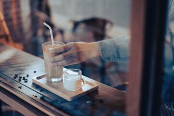 A hand holding a tall glass of iced latte coffee with milk on a wooden bar over a cafe glass window...
