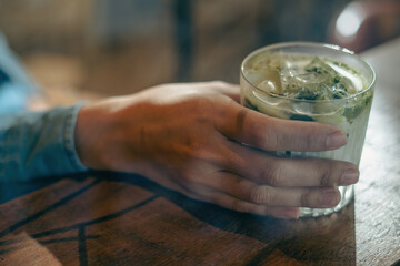 Hand on a glass of Iced matcha latte green tea with milk on a wooden bar over a cafe glass window reflex at a Cafe coffee shop. Cold brew refreshment summer drink with copy and space. Selective focus