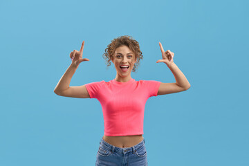 Front view of young slim girl with curly hair standing, raising fingers. Pretty blonde female smiling with open mouth, holding hands up. Isolated on blue studio background.