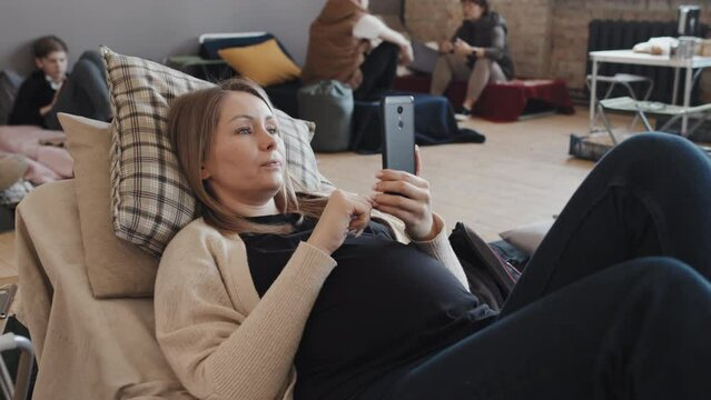Medium long of blonde Caucasian pregnant woman using smartphone, video calling lying on mattress in refugee shelter at daytime