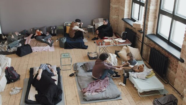 High angle view of diverse refugees lying and sitting on sleeping places, talking, using gadgets, reading books and sleeping at daytime