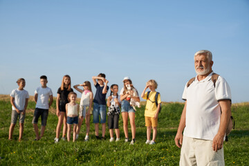 Front view of old male with gray hair and beard looking forward with children outside. Group of tourists having rest, enjoying landscape in mountains. Concept of beauty of nature.