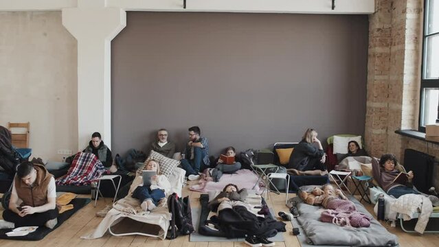 Long view of children, women and men on cots and mattresses in indoor refugee camp at daytime, talking, reading, using gadgets