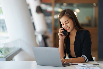 Happy secretary Asian business woman charming sitting smiling and talking on the smartphone in the office.