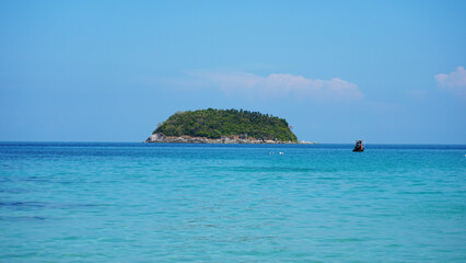 A lonely island in the sea. A Thai fishing boat floats nearby. Clear blue water, snow-white sand. Small clouds in the sea. Palm trees and trees grow on the island. Beautiful Kata beach, Phuket