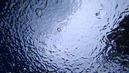 View from under the water to the sky and rain. Drops fall on the surface of the water create water circles. Gray clouds and blue sky are visible. Dark green highlights of trees. An unusual look