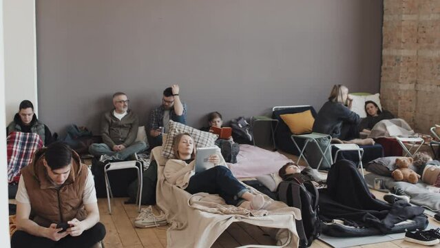 View of children, women and men on cots and mattresses in indoor refugee camp at daytime, talking, reading, using gadgets