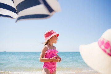 family with child relax on beach by sea in summer. girl in pink swimsuit and panama on background of turquoise sea, umbrella and woman's hat