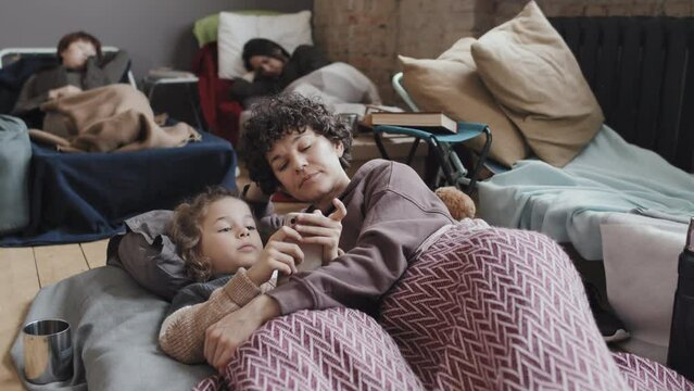 Medium long of curly woman hugging her adorable Caucasian son, talking and looking at smartphone, lying on mattress in flee camp at daytime