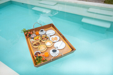 Floating Breakfast tray in swimming pool at luxury hotel or tropical resort villa, fruits, croissant, coffee, and orange juice. Exotic summer, relaxation, tropical travel and vacation concept