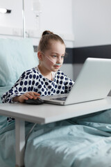 Smiling sick little girl playing games on computer laptop while in pediatric clinic recovery ward room. appy ill little child enjoying video games on laptop while resting in hospital bed.