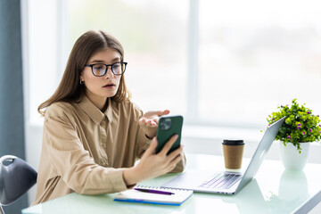 Young smiling fun successful employee business woman use hold mobile phone sit work at workplace white desk with laptop at office indoors. Achievement career concept.