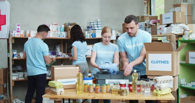 Caucasian male and female activists in blue t-shirt working together at warehouse of charitable organization. Team of volunteers sorting provision on shelves and packing in cardboard boxes.