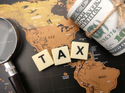 Magnifying glass and banknotes with the word TAX on a world map background.