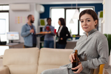 Portrait of young woman holding beer bottle after hours in office, celebrating with workmates. Happy person enjoying leisure activity and party after work, having alcoholic drinks.