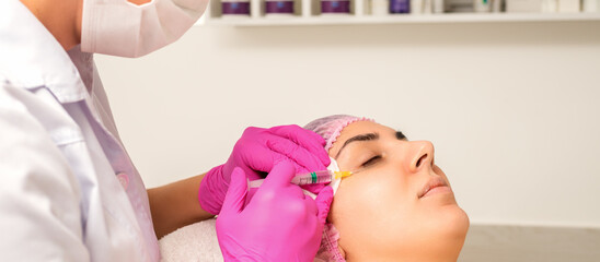 The young white woman is getting rejuvenating facial injections with hyaluronic acid on the eye in a beauty clinic