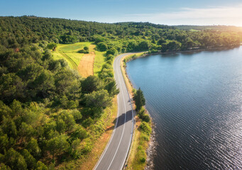 Aerial view of road near blue sea, forest at sunset in summer. Travel in Croatia. Top view of beautiful road, green trees, hills in spring. Colorful landscape with highway and sea coast. Road trip	