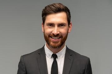 Isolated shot of young handsome male with beard and trendy hairdo, wearing formal clothes posing with toothy smile in studio against grey background