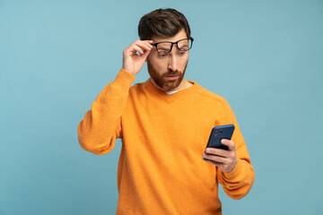 Mobile connection. Portrait of shocked astonished man reading post on social network using cell phone, chatting looking surprised. Indoor studio shot isolated on blue background - obrazy, fototapety, plakaty