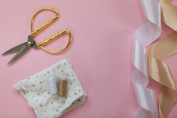 The layout of sewing accessories and fabric on a pink background. Sewing threads, needles, fabric, buttons and scissors in gold and white. top view, flatlay with space for text