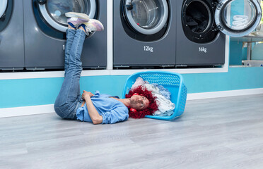 A young Latina woman with red afro hair lying on the floor with her head in the basket in a blue automatic laundromat listening to music with red headphones while waiting for the laundry to be done.