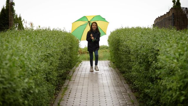 Cute little girl running under umbrella in rainy spring day along green bushes towards camera