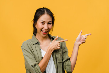 Portrait of young Asian lady smiling with cheerful expression, shows something amazing at blank space in casual cloth and looking at camera isolated over yellow background. Facial expression concept.