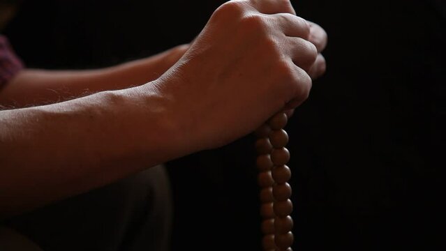 man sorting with two hands a round wooden rosary on a black background close-up. A dim light falls from above