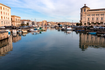Naklejka na ściany i meble View of fishing boats moored in historic inner harbor in the city of Syracuse (Siracusa), Sicily, Italy, Europe, EU. Port connecting Syracuse with Ortygia Island. Reflection in water on a sunny day