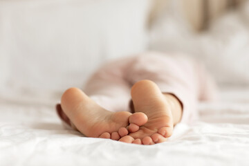 View on kid bare feet on white bed. Bedroom lit with morning light. Child sleeps in pajamas comfort
