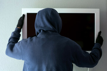 A man in a hooded sweatshirt and black gloves removes the TV from the wall. The theme of theft and...