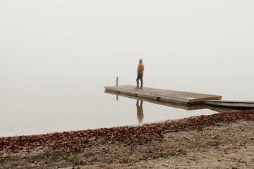 Fototapeta na wymiar A man is standing on a wooden pier at lake Bohinj in Slovenia. He is facing the distance. The background is foggy. Moody hazy winter landscape. He is thinking, and contemplating. Copy space. 