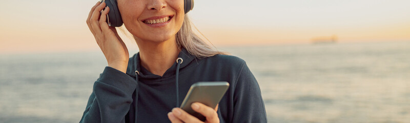Mature happy woman standing on the beach and listening to music on wireless headphones on background of sky at sunset and sea