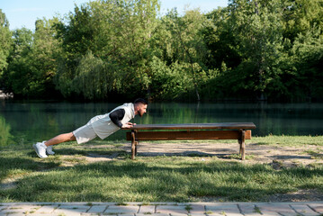 Young brunette man do morning exercises in city park pushing his body up from parks' bench