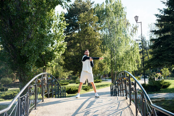Young brunette man do morning exercises in city park standing on bridge and wave his hands